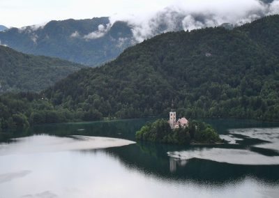 A small island with a church sits in the middle of a calm Slovenian lake, surrounded by lush green forested hills and misty clouds in the background.
