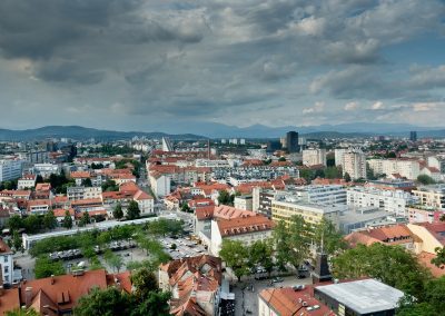 A panoramic view of a Slovenian city with red-roofed buildings, modern high-rises, and tree-lined streets under a partly cloudy sky, with mountains visible in the distant background.