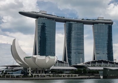 The image shows Singapore's iconic Marina Bay Sands, featuring three tall glass towers connected at the top by a large rooftop structure, with the lotus-shaped ArtScience Museum in the foreground beside the waterfront.