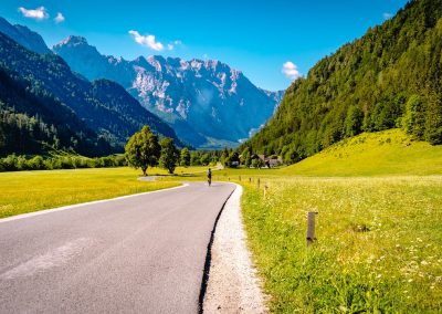 A winding road curves through a green meadow in Slovenia, bordered by forested hills and tall mountains under a bright blue sky. A person rides a bicycle along the road towards distant alpine peaks.