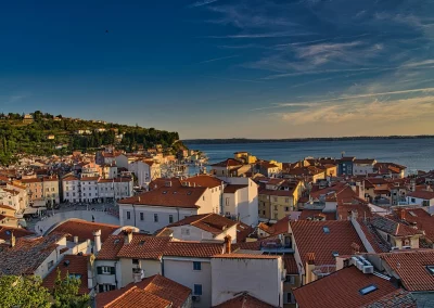 A coastal town in Slovenia with red-tiled rooftops and white buildings, nestled beside a calm blue sea under a clear sky, with green hills rising in the background.