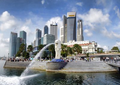 The iconic Merlion statue in Singapore spouts water into Marina Bay, as groups of people gather nearby and modern Singapore skyscrapers rise in the background under a partly cloudy sky.