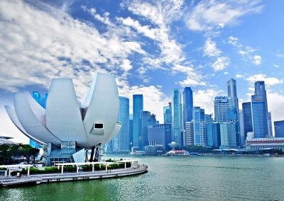 A futuristic white lotus-shaped building by the Singapore waterfront, with the city’s modern skyscraper skyline in the background under a partly cloudy blue sky.