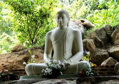 A white statue of Buddha sits cross-legged in meditation on a stone platform, surrounded by green trees, rocks, and floral offerings at the base, reflecting the serene beauty often found at Buddhist sites in Sri Lanka.