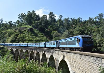 A blue passenger train crosses a stone arched bridge in Sri Lanka, surrounded by lush green hills and trees under a clear blue sky.