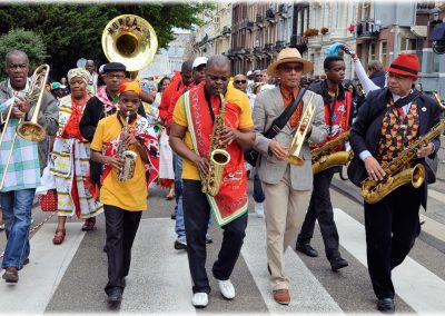 A lively street parade in Suriname shows musicians playing saxophones, a sousaphone, and other brass instruments, dressed in colourful clothes and hats, walking down a city street with onlookers in the background.