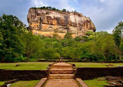 A large, flat-topped rock formation rises above lush green trees and gardens in Sri Lanka, with a stone pathway leading towards it under a partly cloudy sky.