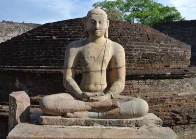 A stone statue of a seated Buddha in a meditative pose, set against the ancient brick ruins and lush greenery of Sri Lanka under a partly cloudy sky.