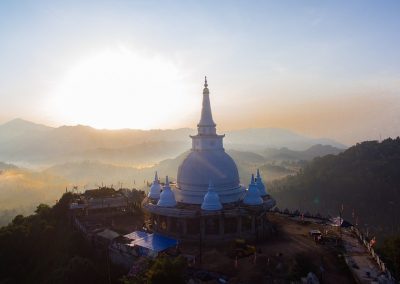 A large white stupa stands on a hilltop in Sri Lanka, surrounded by trees, with mountains and valleys in the background, bathed in soft sunrise light and mist.