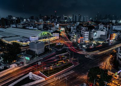 A night-time cityscape showing busy streets with bright light trails from moving vehicles, illuminated buildings, and a large, well-lit railway station among high-rise structures under a dark sky.