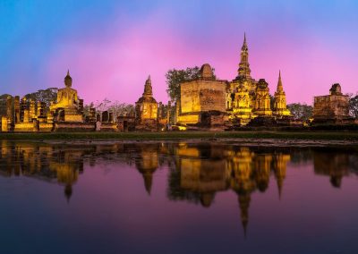 Ancient temple ruins and Buddha statues are reflected in a calm pond at sunset, with a pink and purple sky in the background. The scene is tranquil and illuminated by soft, golden lighting.