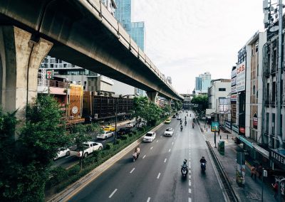 A city street with several lanes of traffic, including cars and motorbikes, runs beneath an elevated railway track. Tall buildings line both sides of the road, and greenery separates the lanes. The sky is partly cloudy.
