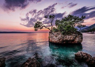 A small rocky island with green trees sits in clear, calm water at sunset, with colourful clouds and a mountain in the background. The sky is pink, purple, and orange, reflecting on the water’s surface.
