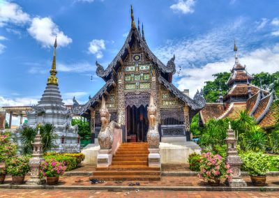 Ornate Buddhist temple with detailed carvings, colourful mosaics, and statues at the entrance, surrounded by lush green plants and a bright blue sky with white clouds. Shoes are placed on the ground by the steps.