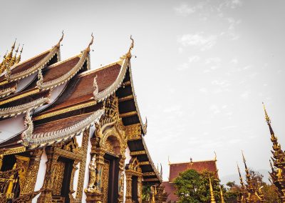 Ornate golden Thai temple roofs and spires with intricate carvings and red accents, set against a pale sky. Some greenery and distant mountains are visible in the background.
